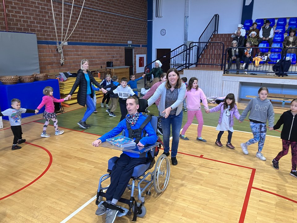 Učenci plešejo v krogu, uporabnik na vozičku ter spremljevalka pa sta v sredini kroga